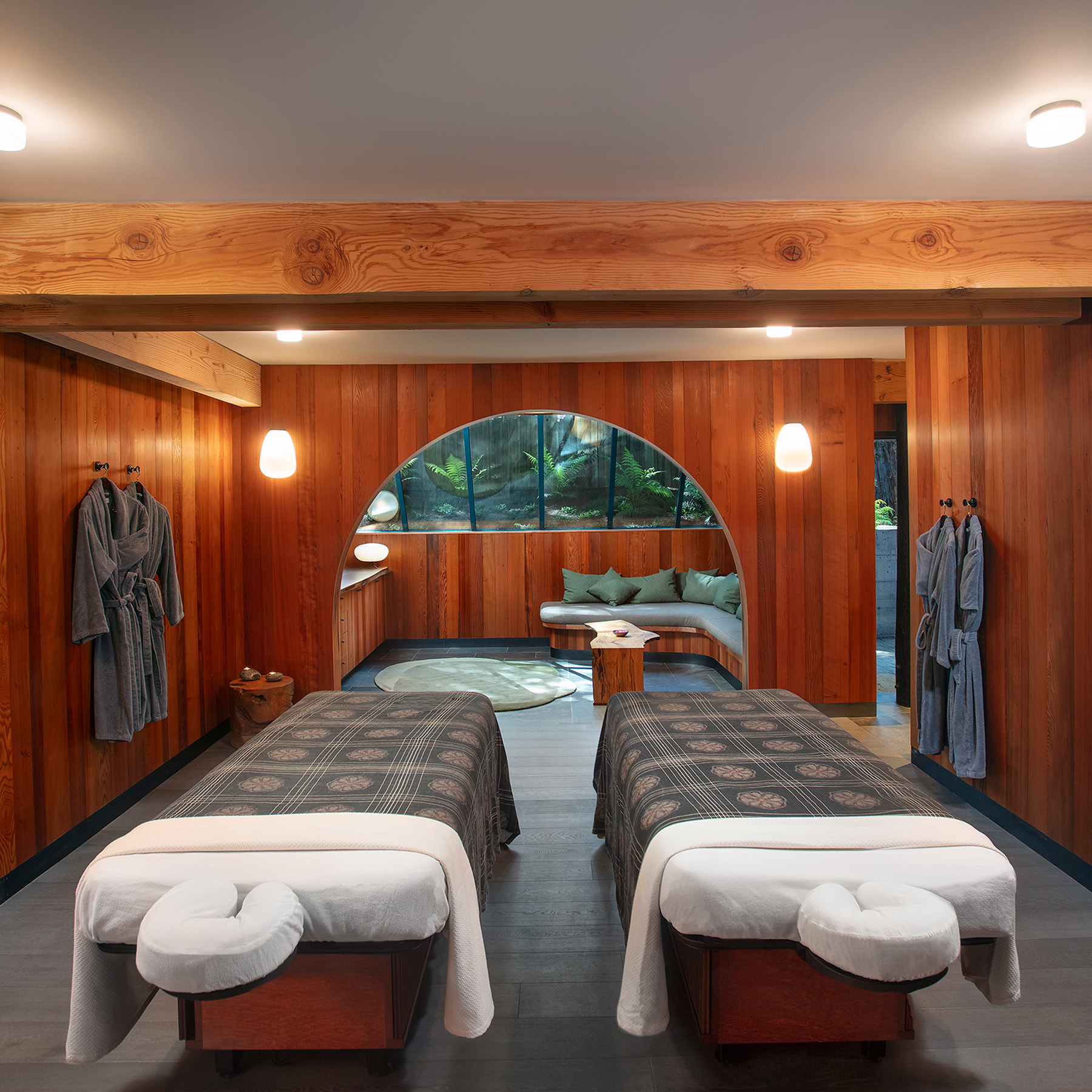 Spa treatment room with two massage tables, wooden paneled walls, robes on hooks, and warm lighting in a serene space.